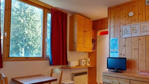 Une cuisine de taille réduite avec des meubles en bois, une table à manger, une chaise, un téléviseur et une fenêtre ornée d ’un rideau rouge.
