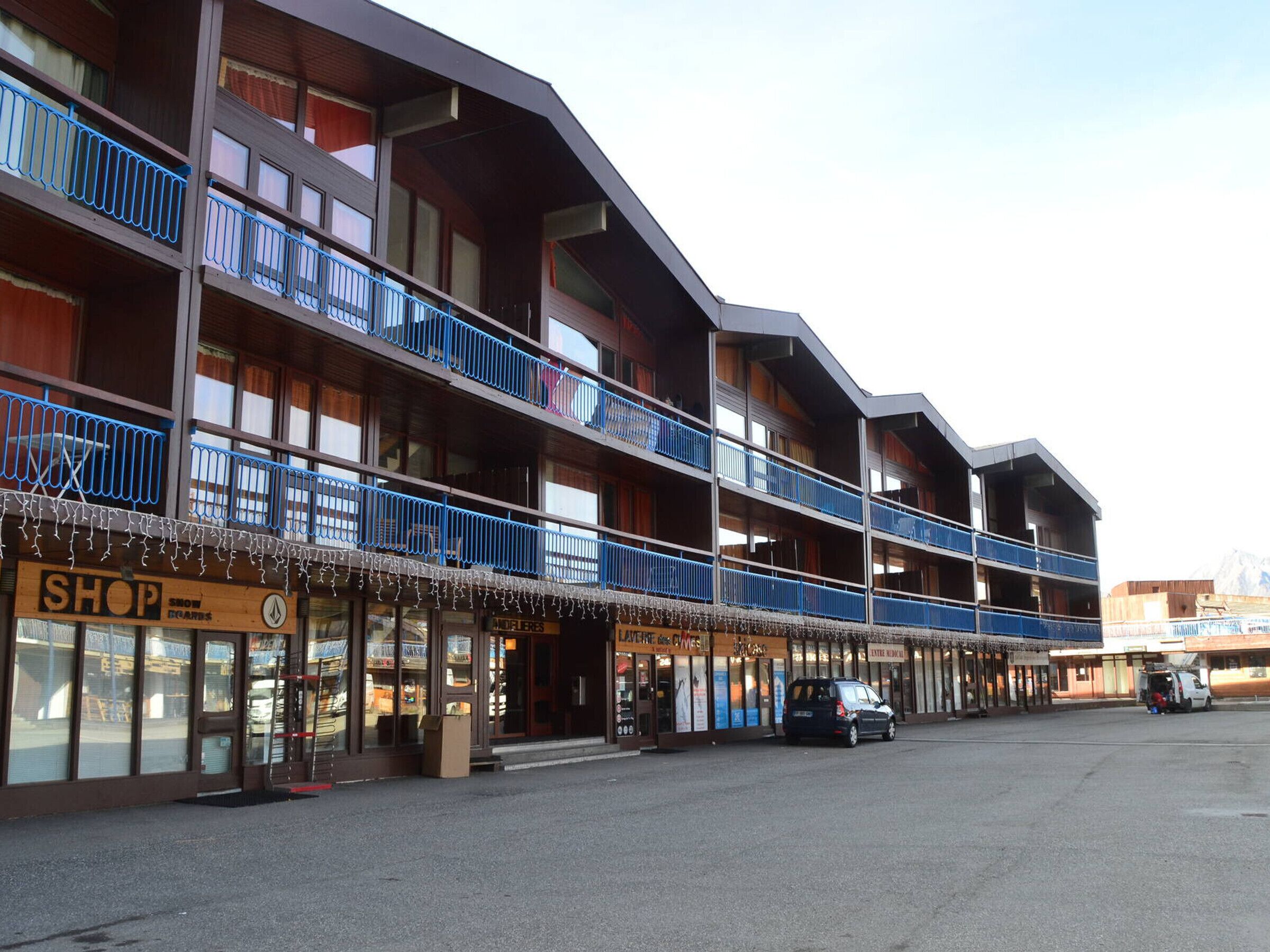 A multi-story building with balconies, a shop on the ground floor, and a parking area.