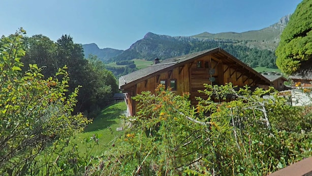 Una baita di legno con tetto spiovente, circondata da vegetazione lussureggiante e montagne sullo sfondo.