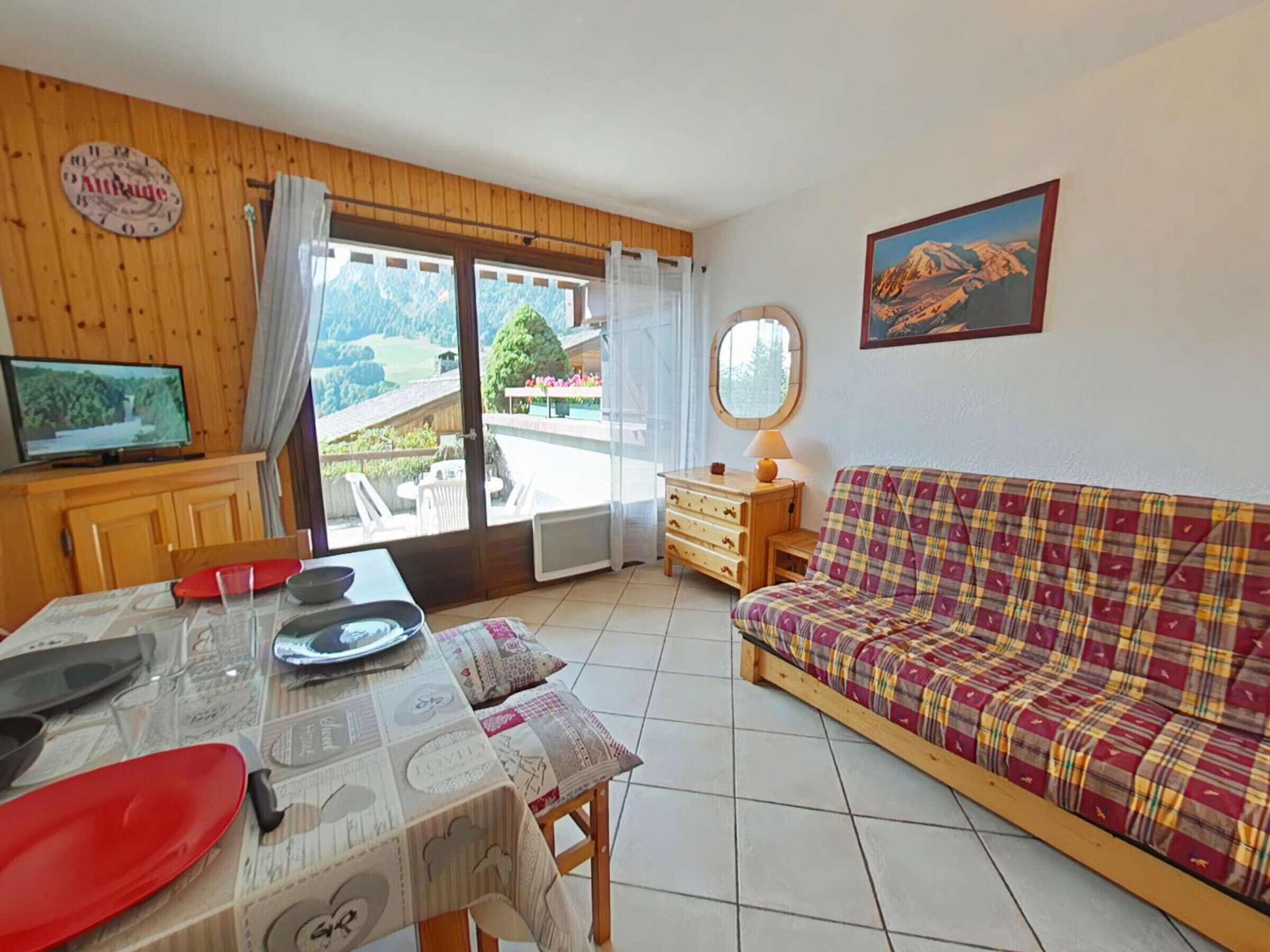 A cozy living room with a wooden table, a plaid sofa, a television, and a scenic view through the glass door.