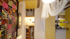 Une chambre avec une couverture colorée, un lit avec du linge de lit blanc et un ciel de lit blanc. Une porte en bois donne accès à une salle de bain équipée d’un miroir et d’un porte-serviettes.