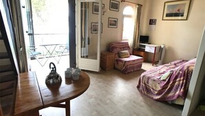 A cozy room with a bed, a desk, a chair, and a table. There is a window with curtains and a door leading to an outdoor area.