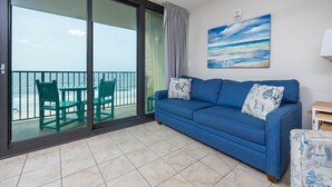 A balcony with a view of the sea, a blue sofa with patterned cushions, and a painting on the wall.