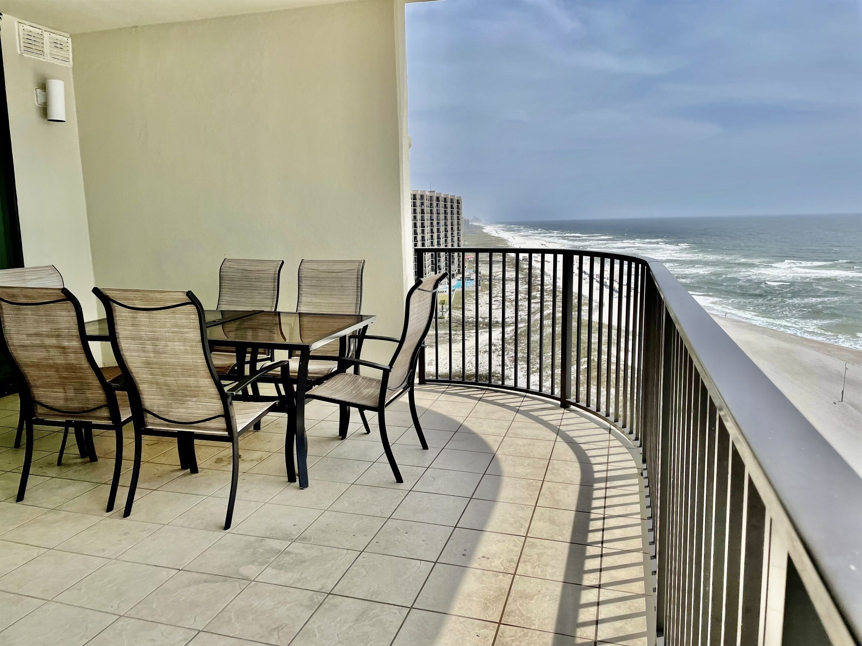 A balcony with a glass table and chairs, overlooking a beach with waves and a distant building.