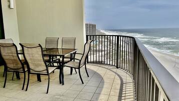 A balcony with a glass table and chairs, overlooking a beach with waves and a distant building.