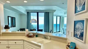 A kitchen with a large window overlooking the ocean, white cabinets, and a countertop with a basket of towels and a bottle of dish soap.
