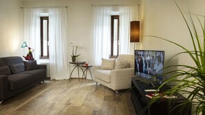 A living room with a dark sofa, a light-colored armchair, a small table with a potted plant, and a TV on a stand. - Welcome to Tabata apartment in the hearth of Florence (Firenze)