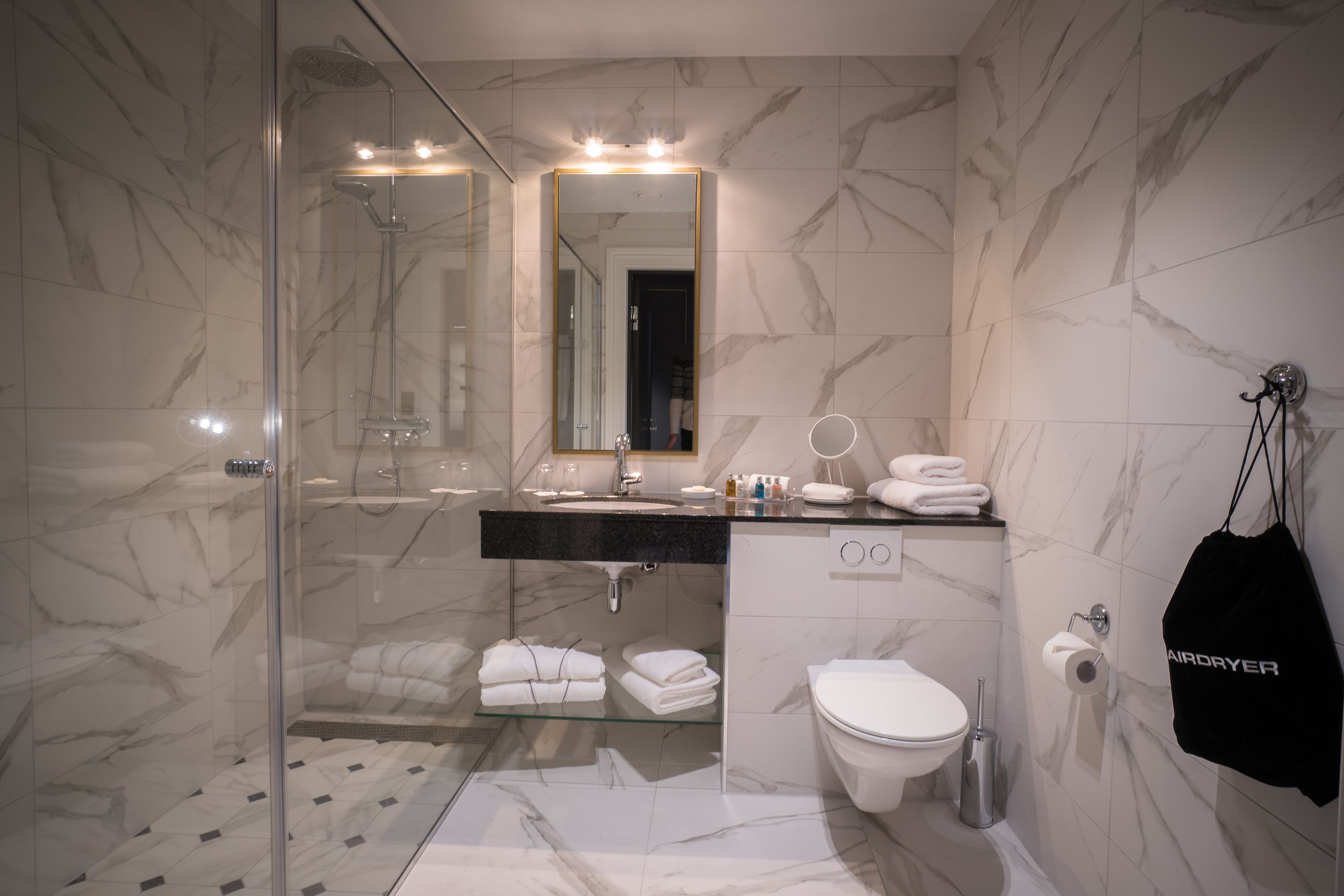 Ein modernes Badezimmer mit einer Duschkabine aus Glas, einem marmorverlegten Boden, einem schwarzen Waschtisch und einer weißen Toilette. Ein Handtuchhalter mit Handtüchern ist ebenfalls vorhanden.
