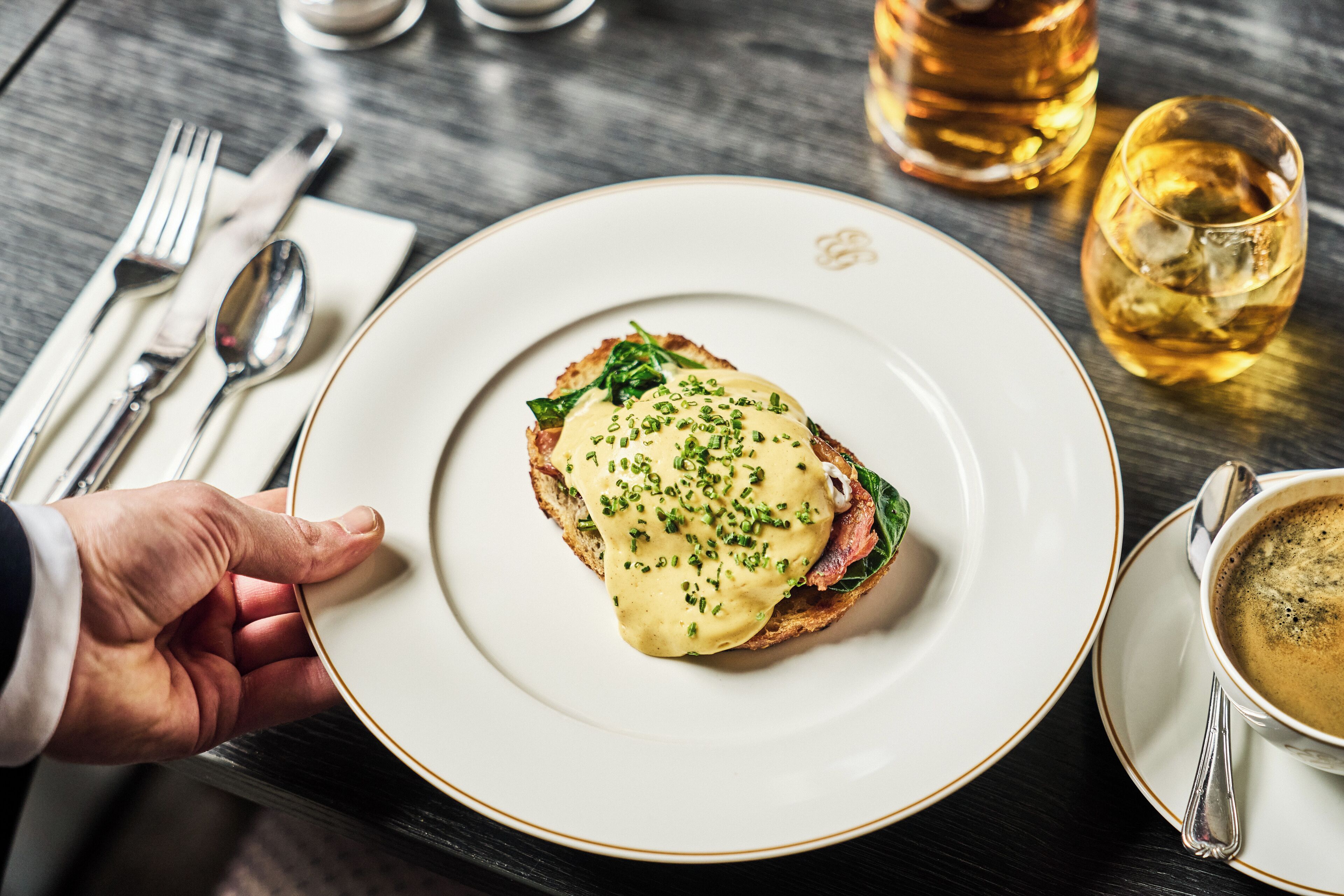 Un plato de comida con una mano sosteniéndolo, una taza de café y una botella de whisky sobre una mesa.