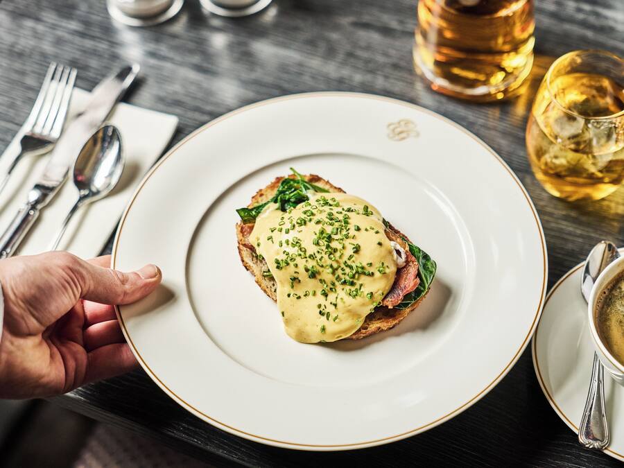 A plate of food with a hand holding it, a cup of coffee, and a bottle of whiskey on a table.