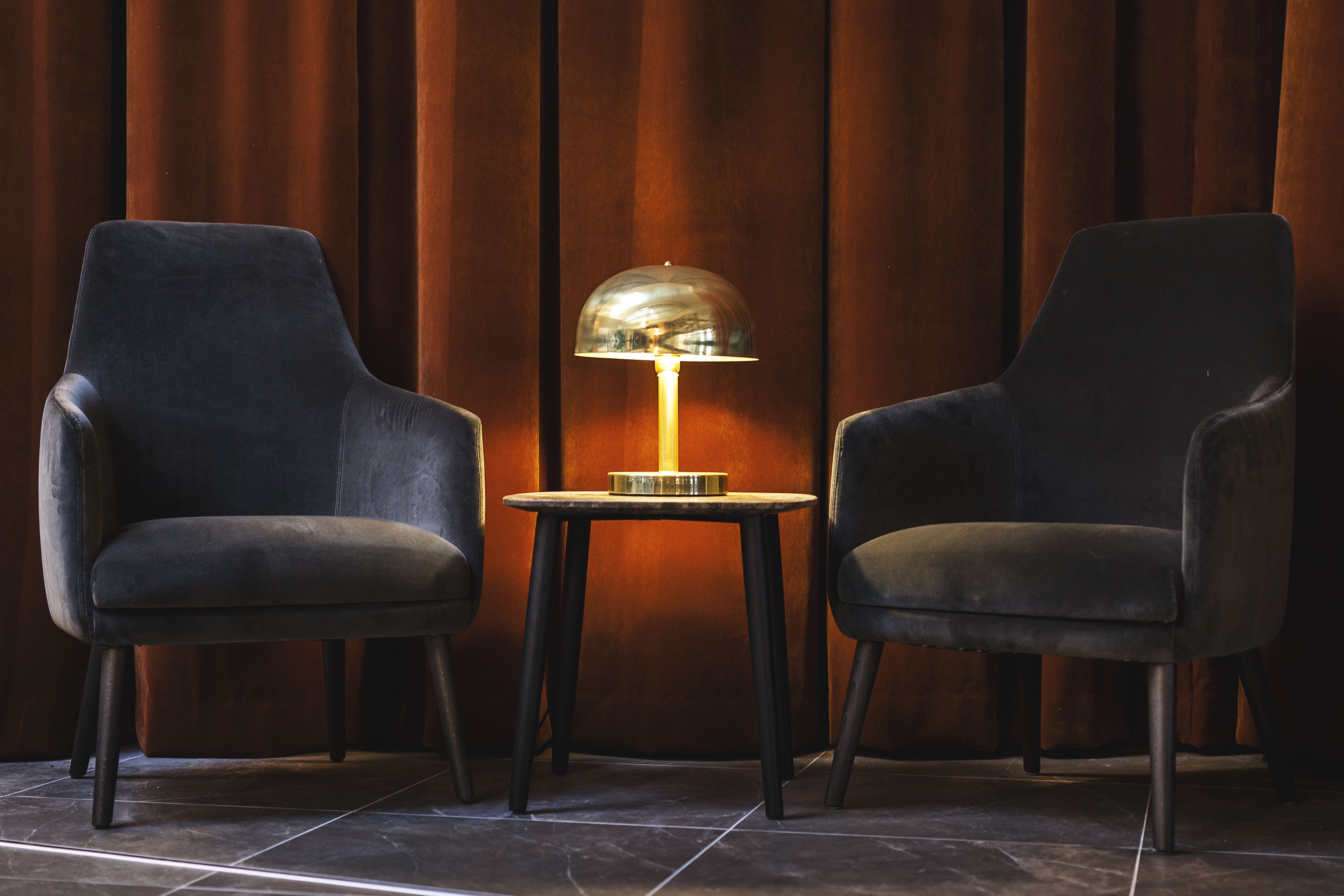 Two black armchairs facing each other with a small round table between them, a lamp on the table, and a wooden panel wall in the background.