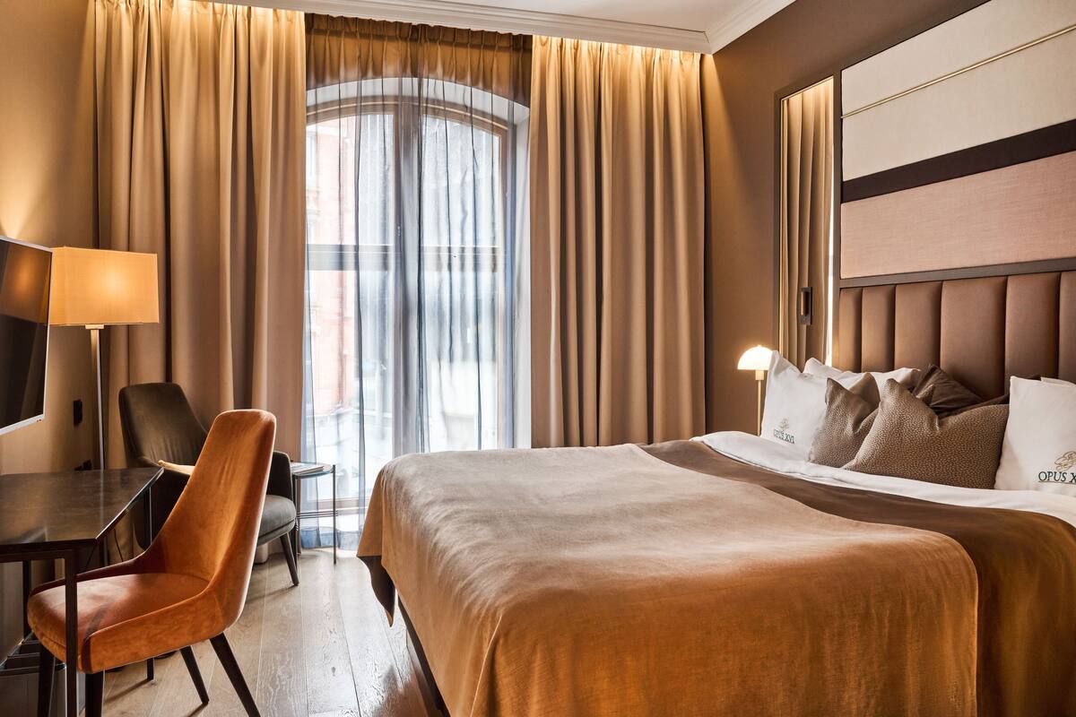 A hotel room with a large bed, a desk with a chair, a TV, and a window with curtains.