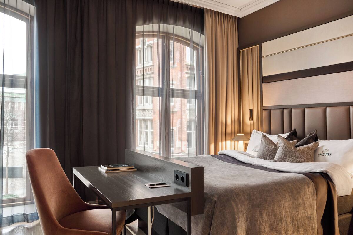 A hotel room with a large bed, a desk with a chair, and a view of a building through the window.