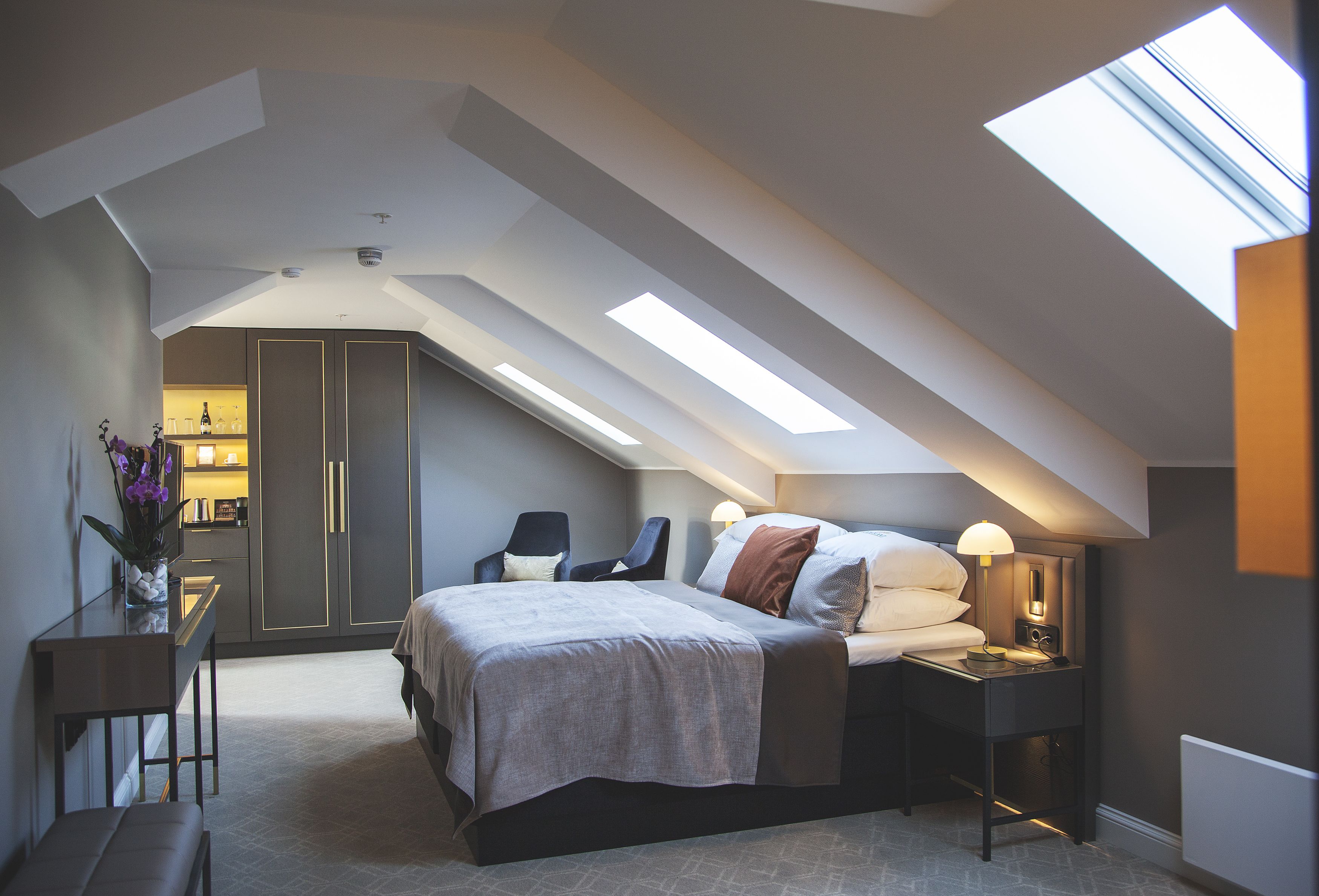 Une chambre moderne avec une lucarne, un lit agrémenté de plusieurs coussins, une table de chevet avec une lampe et un bureau sur lequel est posé un vase de fleurs.