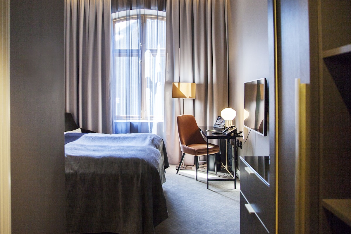 A hotel room with a bed, a desk with a lamp, a chair, and a large window with curtains.