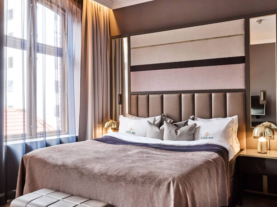 A modern hotel room with a large bed, a nightstand with a lamp, and a large mirror on the wall.