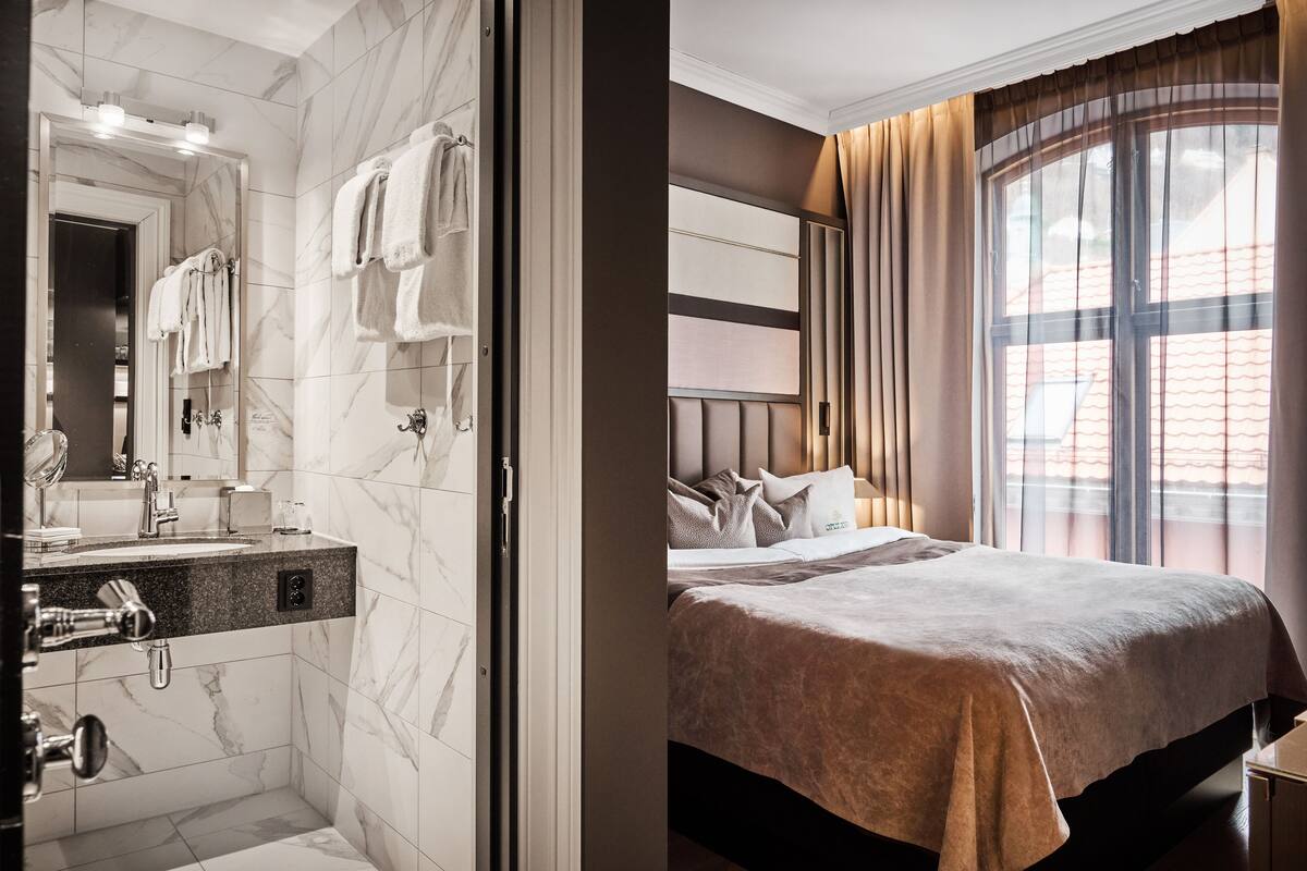A hotel room with a marble bathroom, a bed with a headboard, and a large window with curtains.