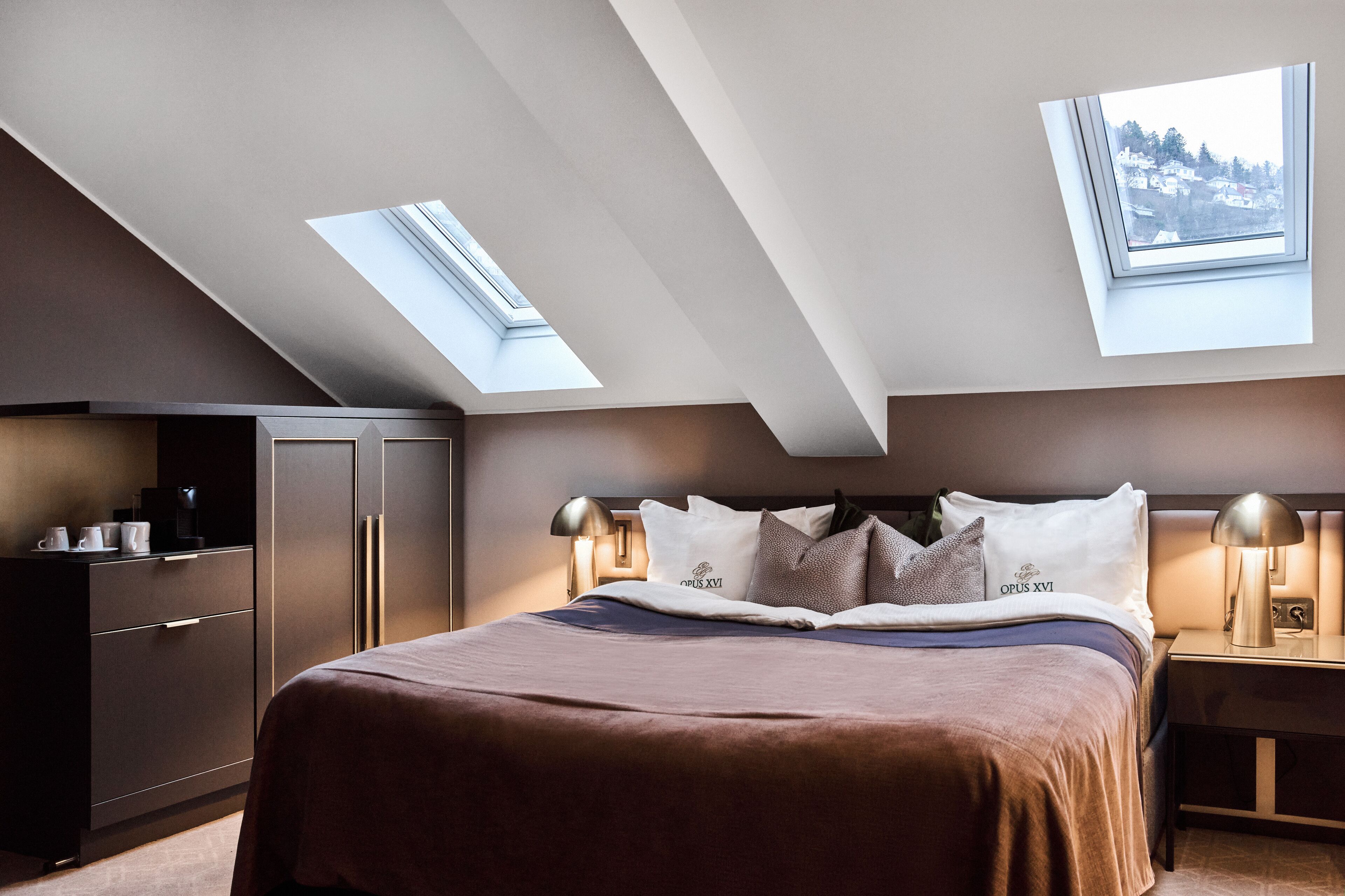 A bedroom with a skylight, a bed with a brown bedspread, a nightstand with a lamp, and a wardrobe.
