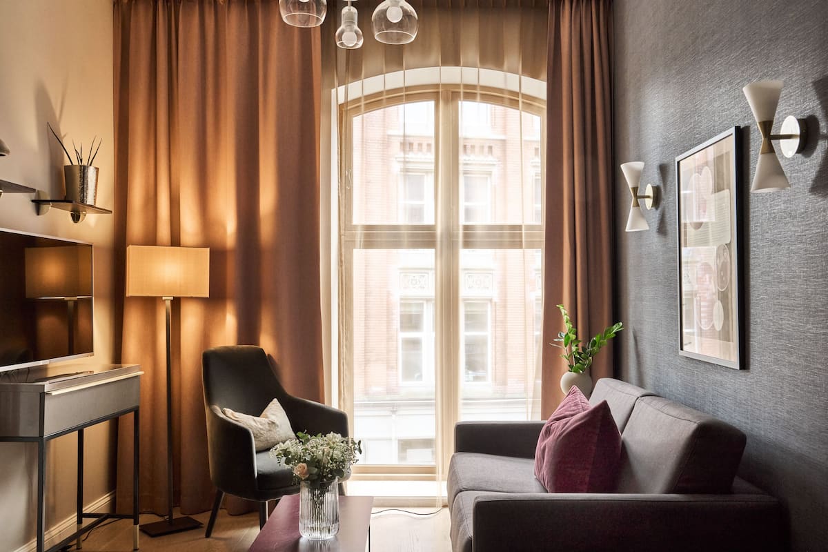 A modern living room with a sofa, armchair, and a desk. A large window with curtains is visible, and there are wall-mounted lights and a framed picture.