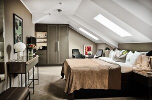 Un dormitorio moderno con techo inclinado, una cama con colcha color beige, un escritorio con lámpara y una silla.