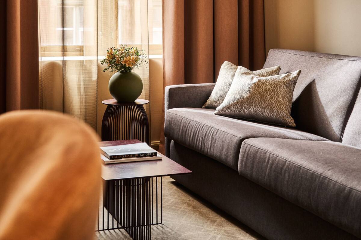 A modern living room with a sofa, a side table with a vase, and a large window with curtains.