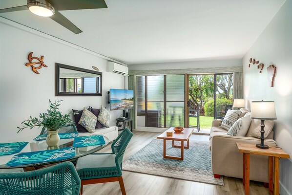 A modern living room with a glass dining table, teal chairs, a beige sofa, and a view of the outdoors through large sliding doors.