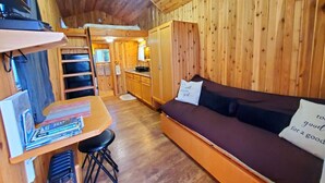 Et interiør af en træhytte med en sofa, et lille bord med bøger, en loftsvifte og et tekøkken.