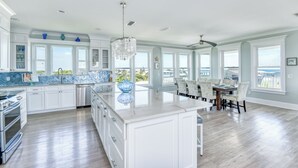 Une cuisine spacieuse avec des meubles blancs et un grand îlot central, un coin repas avec une table et des chaises, et de grandes fenêtres offrant une vue sur l’extérieur.