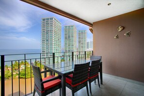 A balcony with a glass table and chairs, overlooking a cityscape and the sea. - Pools, Gym & Restaurant | Beachfront 1BR (Puerto Vallarta)