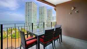 A balcony with a glass table and chairs, overlooking a beach and tall buildings. - Pools, Gym & Restaurant | Beachfront 1BR (Puerto Vallarta)
