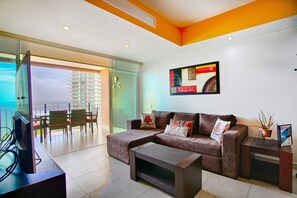 A modern living room with a sofa, coffee table, and a television. There is a balcony with a view of the city skyline. - Pools, Gym & Restaurant | Beachfront 1BR (Puerto Vallarta)