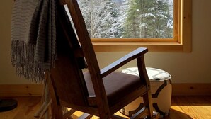 Ein hölzerner Schaukelstuhl mit Decke, vor einem großen Fenster mit Blick auf verschneite Bäume und einen klaren Himmel.
