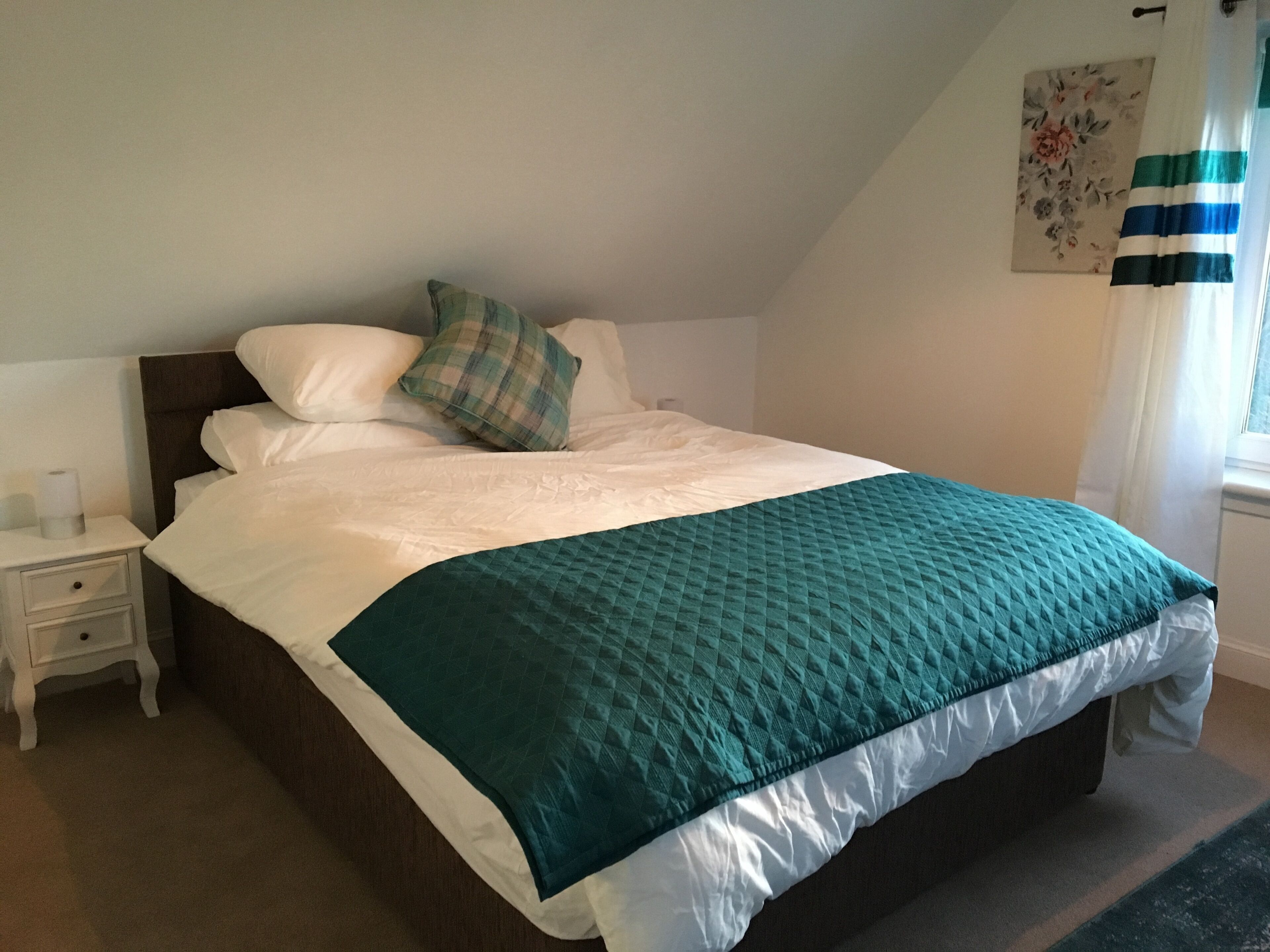 A neatly made bed with a teal quilt, white pillows, and a plaid throw pillow. There is a small bedside table with a lamp and a tissue box. A floral artwork hangs on the wall, and a window with curtains is visible.