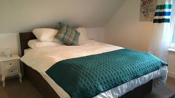 A neatly made bed with a teal quilt, white pillows, and a plaid throw pillow. There is a small bedside table with a lamp and a tissue box. A floral artwork hangs on the wall, and a window with curtains is visible.