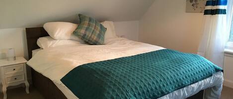 A neatly made bed with a teal quilt, white pillows, and a plaid throw pillow. There is a small bedside table with a lamp and a framed picture on the wall.