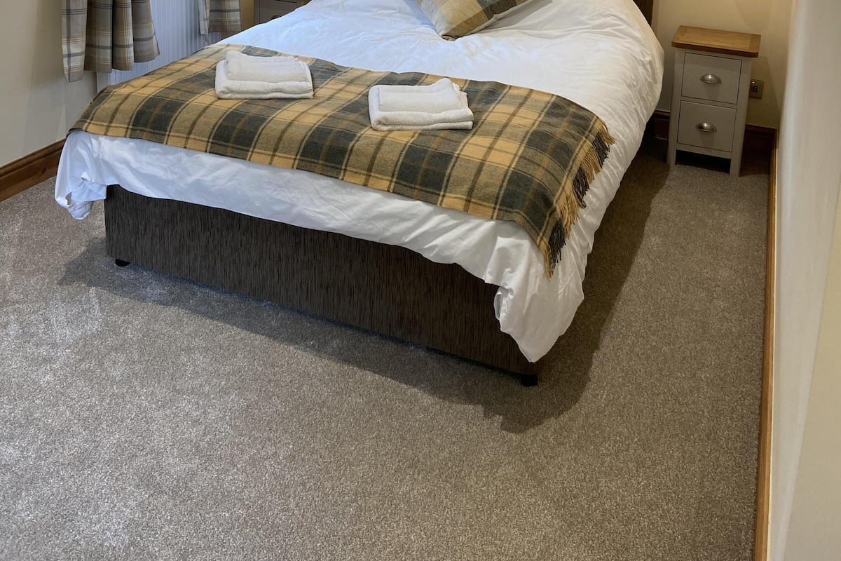 A neatly made bed with a plaid bedspread, two pillows, and two folded towels. There are two bedside tables with lamps, a window with curtains, and a carpeted floor.