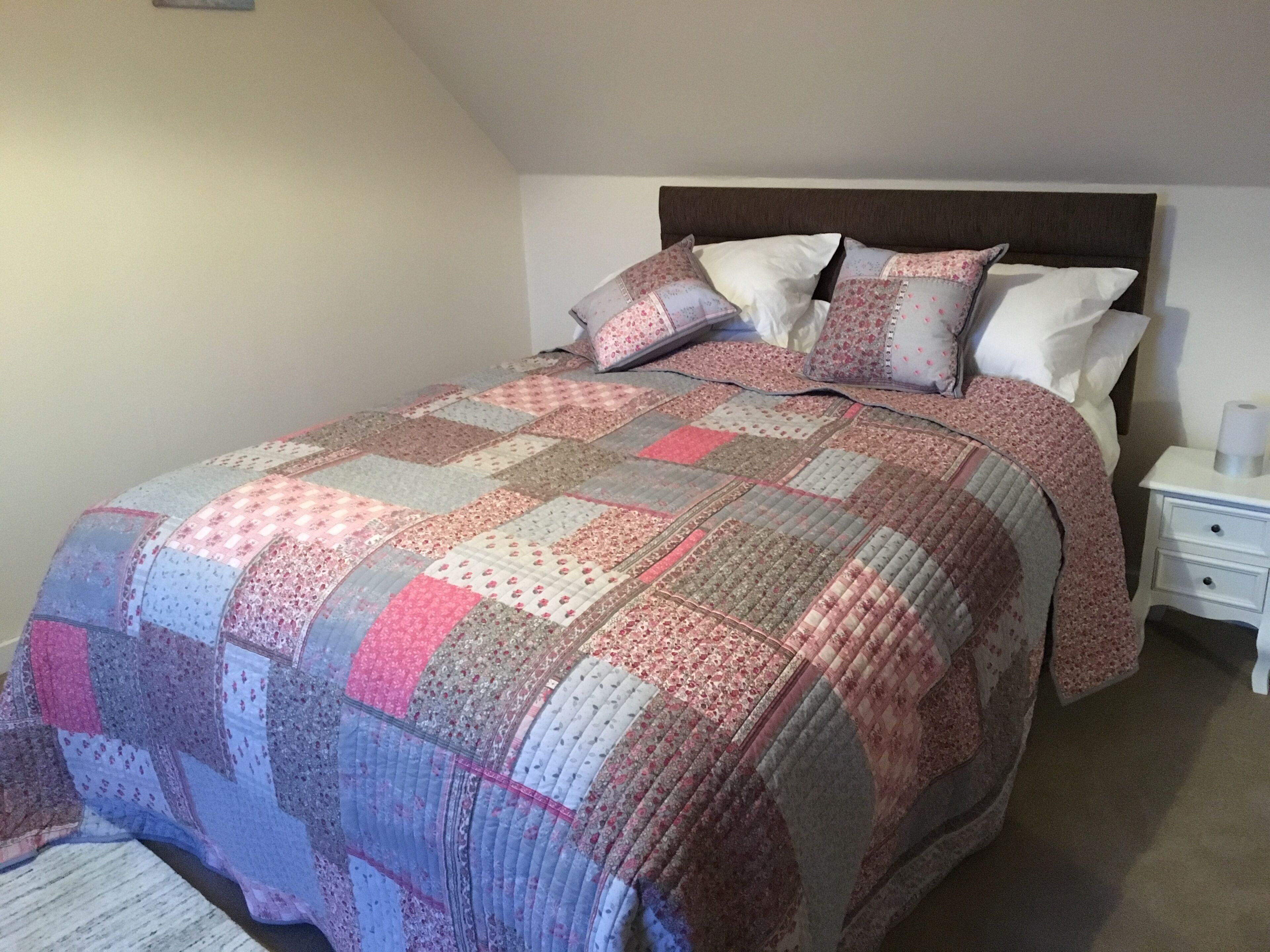 A bedroom with a bed covered in a patchwork quilt, a wooden headboard, and a white nightstand with a lamp.