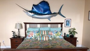 A bedroom with a bed, floral bedding, a wall-mounted fish decoration, a nightstand with a lamp, and a potted plant. - Roomy, Quaint, Studio Loft w/ Bay View - Central SD Location  (San Diego)