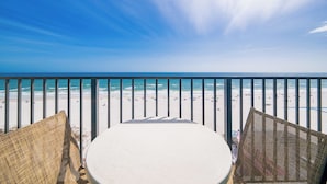 Varanda com cadeiras de vime e uma mesa redonda com vista para uma praia, pessoas e um céu claro.