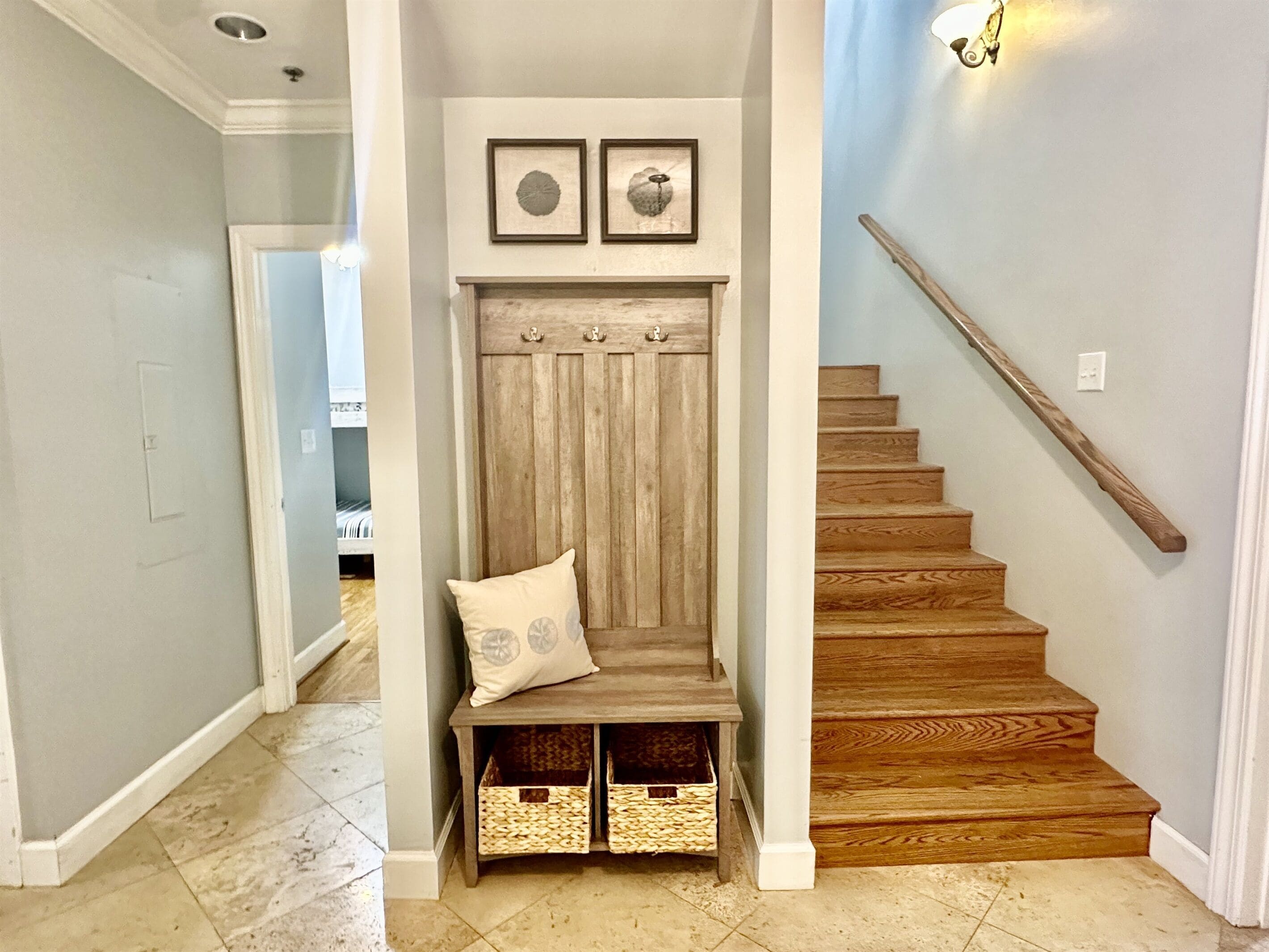 A hallway with a wooden bench, two framed pictures, a staircase with a wooden handrail, and wicker baskets under the bench.
