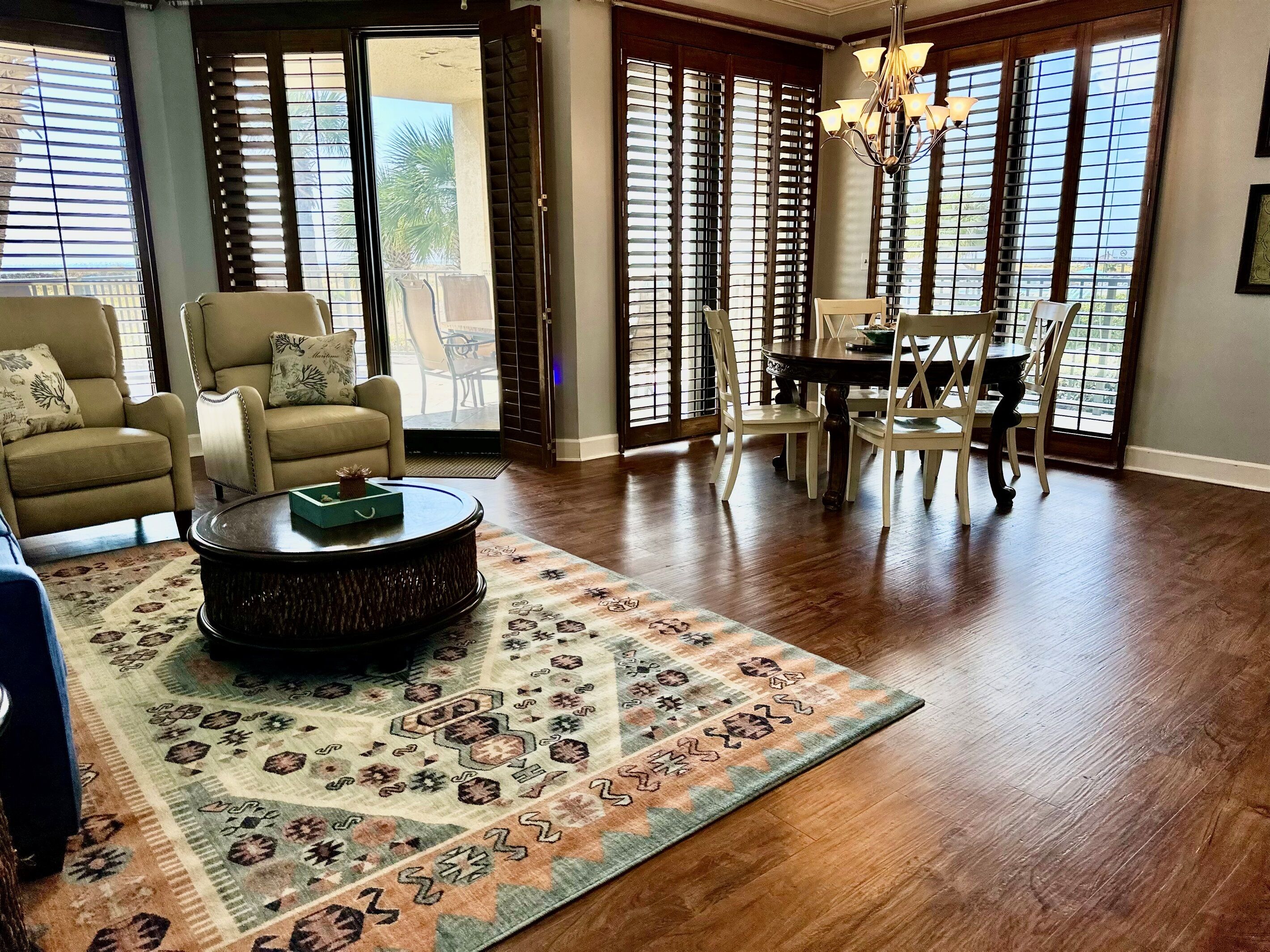 A spacious living room with a large wooden coffee table, a patterned rug, and a dining area with white chairs and a dark wooden table.