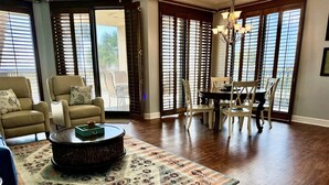 A spacious living room with a large wooden coffee table, a patterned rug, and a dining area with white chairs and a dark wooden table.