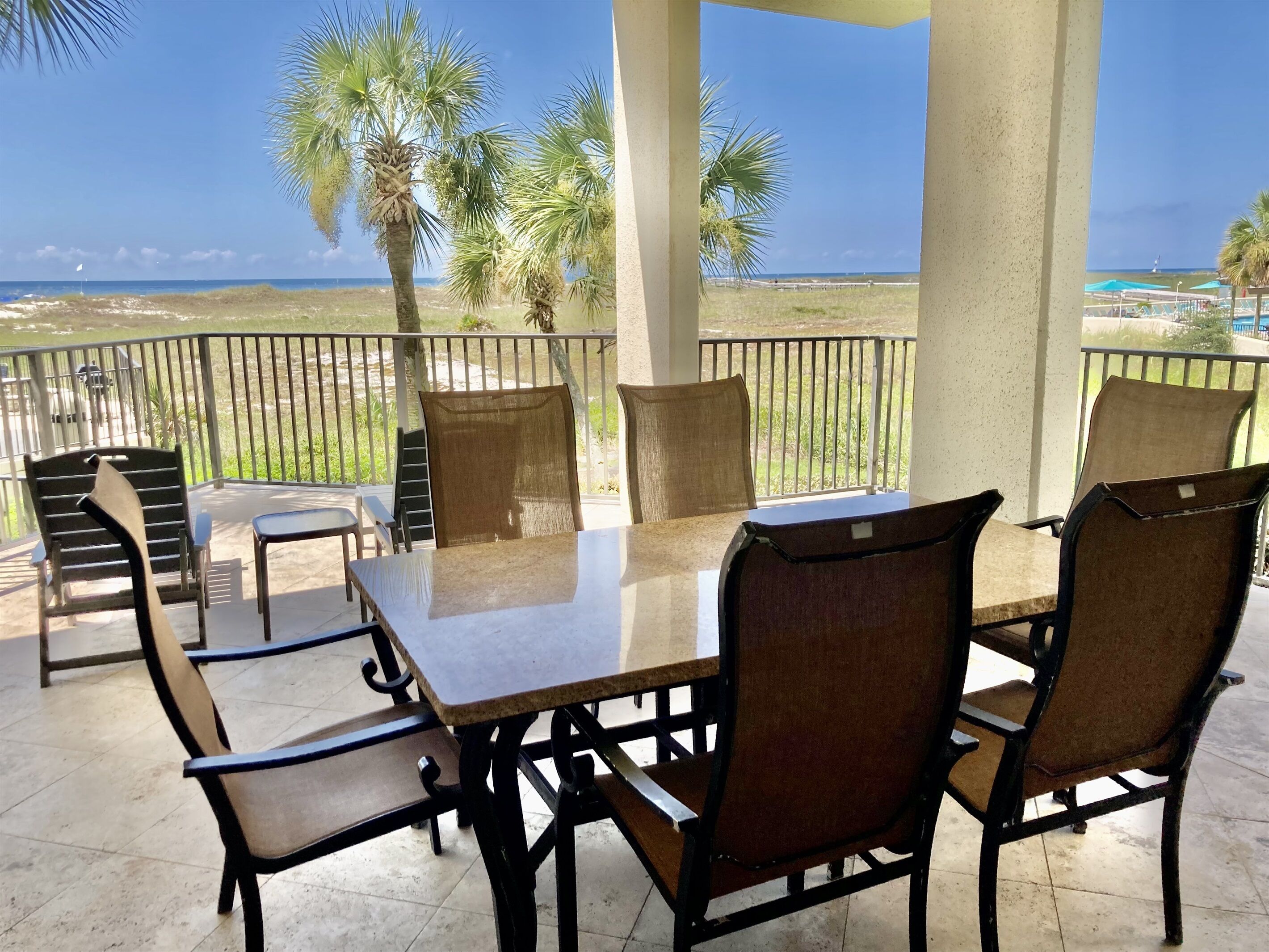 A covered outdoor seating area with a table and chairs, overlooking a beach with palm trees and a clear sky.