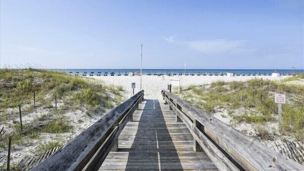 A wooden boardwalk leading to a beach with sand dunes and a clear sky.