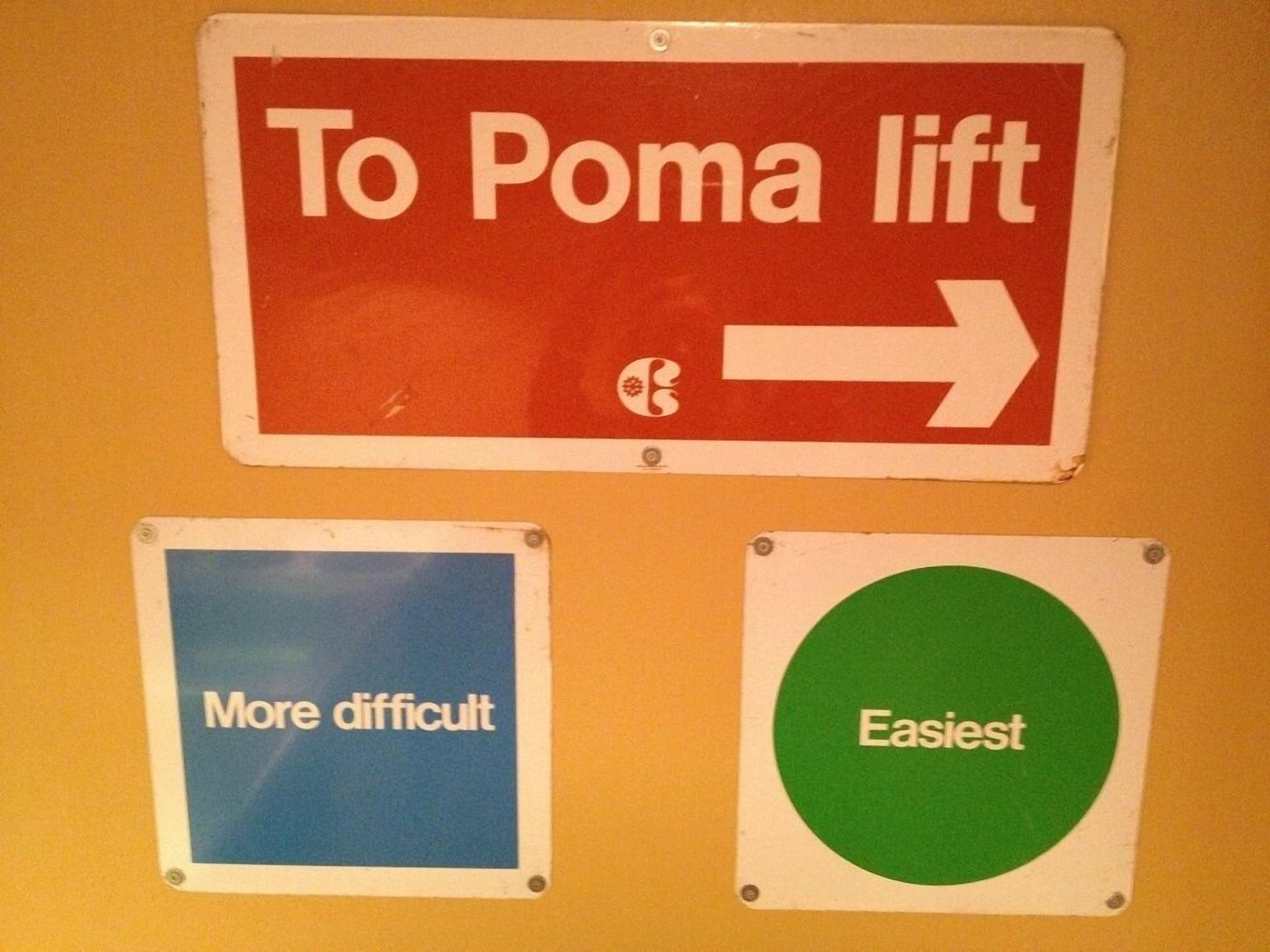 Een muur met drie borden: 'Stoeltjeslift Poma,' 'Moeilijker' en 'Makkelijkst'.