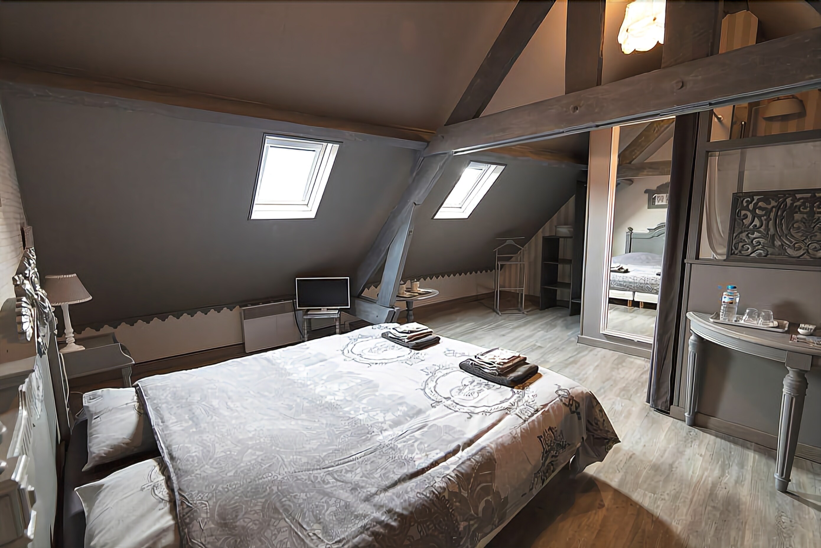 Un dormitorio con techo inclinado, tragaluces, cama con colcha estampada, mesita de noche, lámpara y vista a otra habitación.