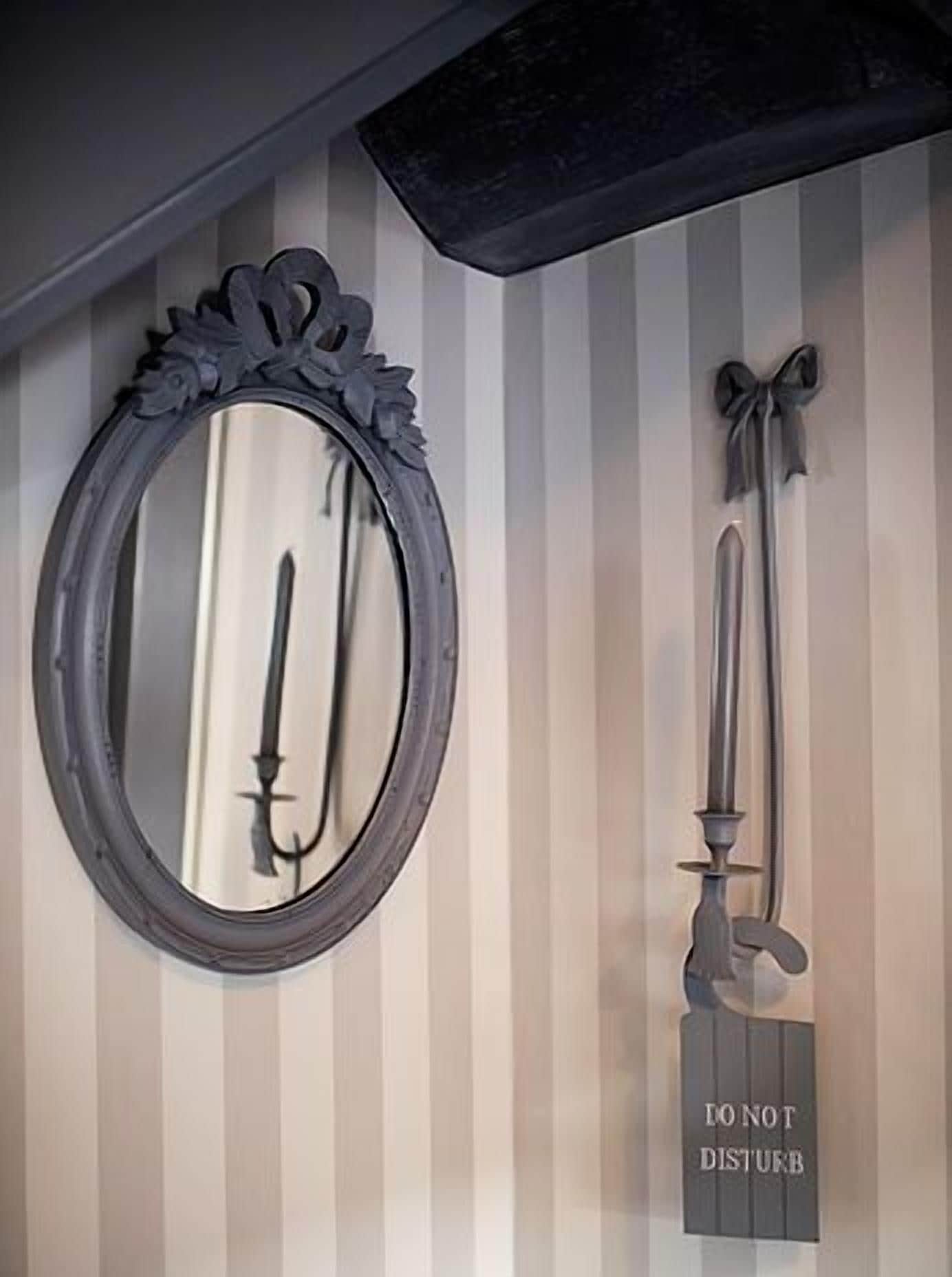 Un miroir avec un cadre décoratif, un chandelier orné d’un nœud et un panneau « Ne pas déranger » sur un mur à rayures.