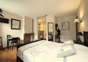 Een slaapkamer met een bed, een bureau, een stoel, een kledingkast, een spiegel en een badkamer door een open deur zichtbaar.