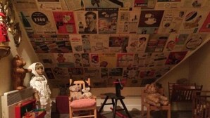 Une pièce avec un plafond en pente recouvert de divers affiches et publicités, une petite chaise en bois, un fauteuil à bascule et un ours en peluche.