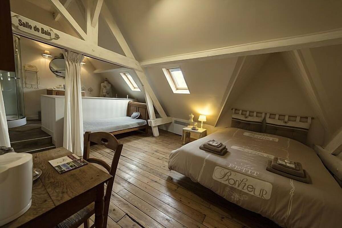 Een gezellig slaapkamer onder de dakpannen met een bed, een klein tafeltje en een stoel. Er is een raam en een deur naar een badkamer.